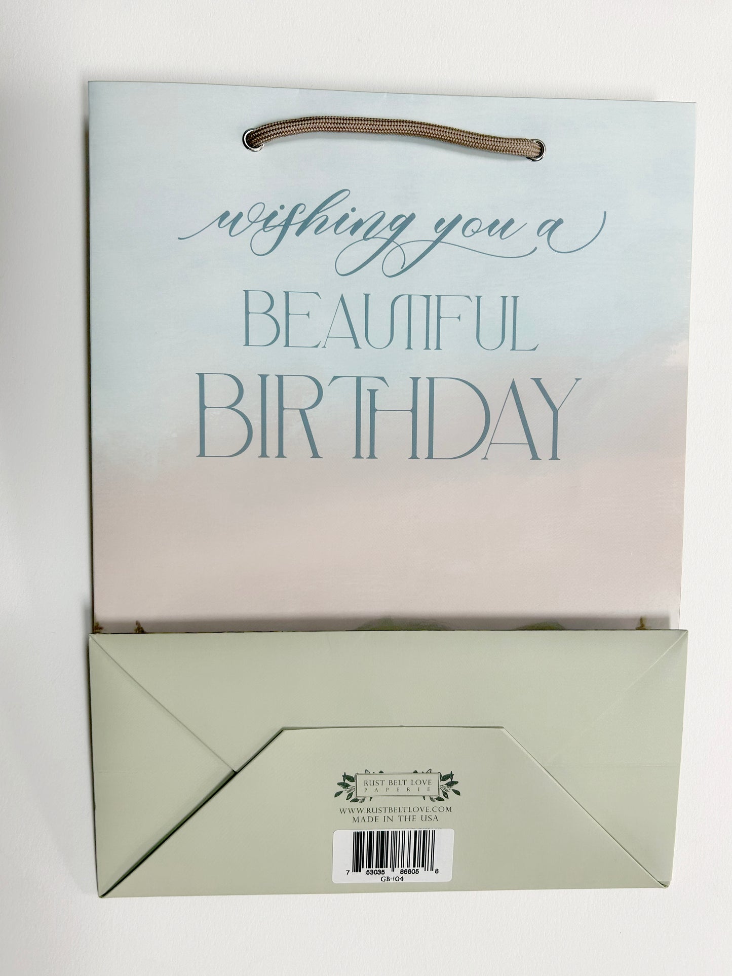 Flatlay photo of Rust Belt Love's birthday bag with mountain and tree illustrations that reads "Wishing you a beautiful birthday". Shows bottom of bag with barcode